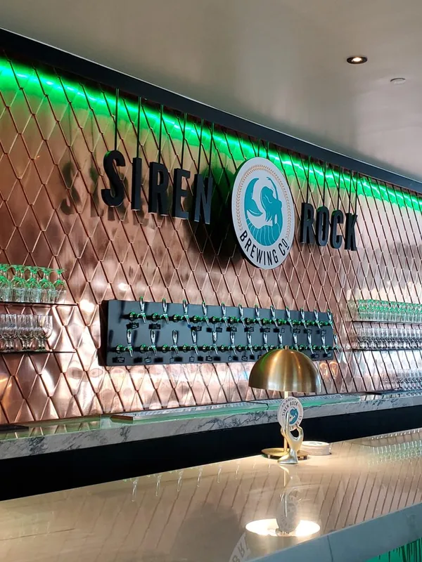 The copper diamond-tile back wall at Siren Rock Brewing Company, the brewery signage and mermaid mark mounted flush, white marble bar in front