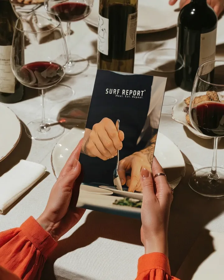 A printed Surf Report menu held open at a set table with wine glasses and bottles.